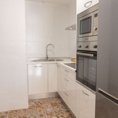 Cocina compacta con muebles blancos, electrodomésticos de acero inoxidable, encimera funcional y suelo de baldosas decorativas.