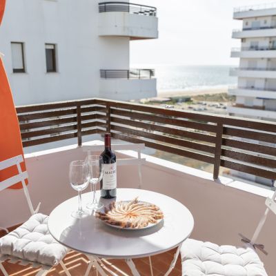 Balcón con mesa y sillas blancas, botella de vino, copas y plato de marisco, con vistas al mar y ambiente relajante.