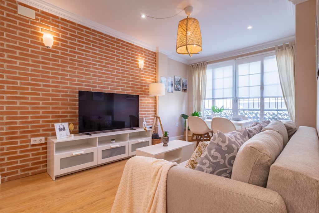 Salón-comedor acogedor con pared de ladrillo visto, sofá en tonos beige con cojines decorativos, televisor sobre mueble blanco y mesa de comedor junto a grandes ventanales con cortinas traslúcidas. Iluminación cálida con lámpara de mimbre y apliques de pared