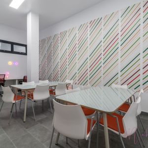 Moderna sala de reuniones y estudio en la Casa de la Juventud con mesas de cristal, sillas blancas y naranjas, mural decorativo a rayas y área de coworking con ordenadores