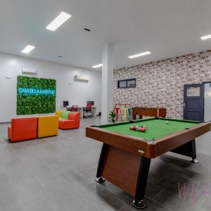 Moderno centro juvenil con mesa de billar, zona de descanso con sofás de colores y un letrero de neón sobre un jardín vertical que dice 'Casa de la Juventud'.