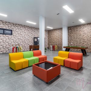 Zona recreativa en un centro juvenil con sofás modulares de colores, mesa de billar, futbolín y taburetes altos. Espacio amplio con paredes de diseño rústico y suelo de baldosa gris, ideal para actividades sociales y de ocio.
