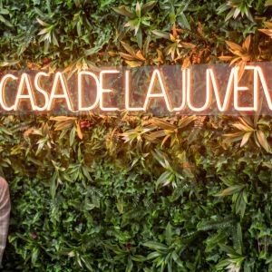 Mujer sonriente posando junto a un letrero de neón con la frase 'Casa de la Juventud', montado sobre un muro verde decorativo con plantas artificiales