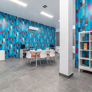 Espacio juvenil moderno con paredes de diseño en tonos azules, estantería con libros, mesas con sillas blancas y escritorios con ordenadores. Ambiente vibrante con decoración colorida y suelo de baldosa gris.