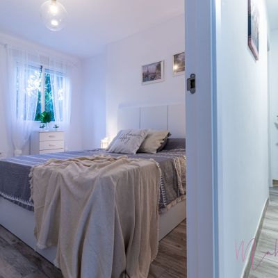 Dormitorio moderno con cama de matrimonio, decoración en tonos neutros, cortinas blancas, aire acondicionado y suelo de madera clara.