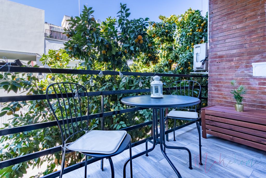 Acogedor balcón con mesa y sillas de hierro, luces decorativas y vistas a árboles frutales en un entorno urbano.