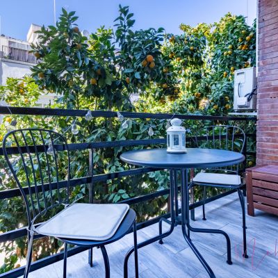 Acogedor balcón con mesa y sillas de hierro, luces decorativas y vistas a árboles frutales en un entorno urbano.