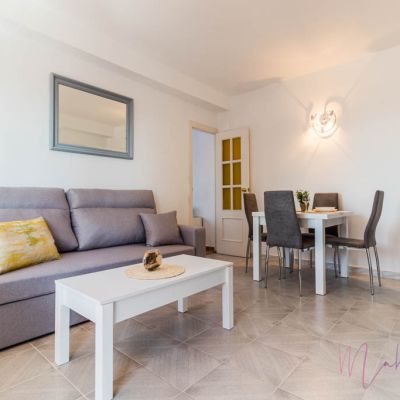 Salón-comedor luminoso con sofá gris, cojín decorativo amarillo, mesa de centro blanca y comedor moderno con sillas tapizadas.