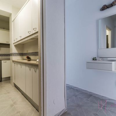 Vista de la cocina con muebles blancos y encimera de madera clara, junto a un recibidor con espejo, estantería flotante y decoración minimalista.
