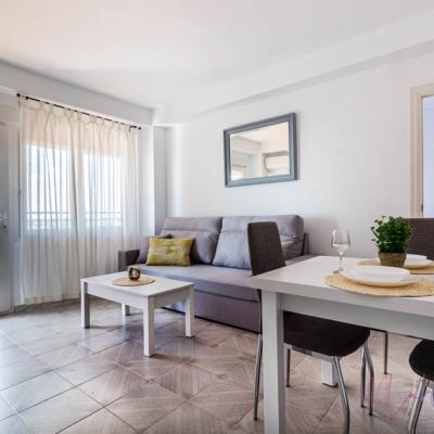 Salón-comedor luminoso con sofá gris, mesa de centro blanca, comedor moderno y acceso a balcón con vistas panorámicas.