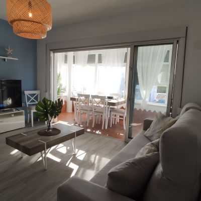 Salón moderno con sofá beige, mesa de centro de madera, televisor y acceso a terraza acristalada con comedor.