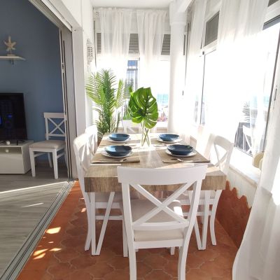 Comedor en terraza acristalada con mesa de madera, sillas blancas, vajilla azul y cortinas blancas translúcidas con vista al exterior.