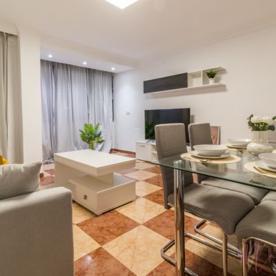 Salón-comedor moderno con sofá gris, mesa de cristal con sillas tapizadas, televisor y decoración en tonos neutros.