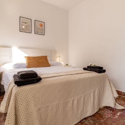 Dormitorio acogedor con cama de matrimonio, ropa de cama en tonos neutros, cojín mostaza, toallas negras y decoración minimalista.