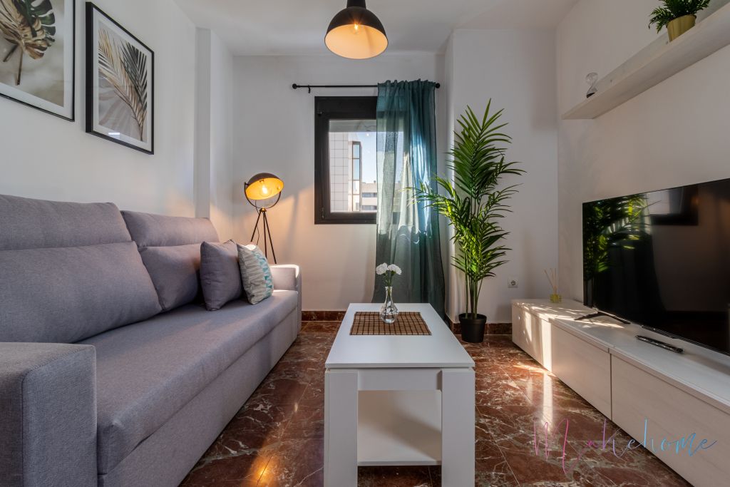 Salón moderno y acogedor con sofá gris, mesa de centro blanca, televisor sobre mueble minimalista y decoración con plantas. Iluminación cálida con lámpara de pie y cortinas en tonos oscuros que contrastan con el suelo de mármol.