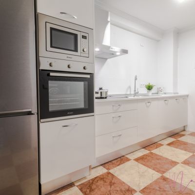 Cocina moderna con muebles blancos, electrodomésticos de acero inoxidable, encimera amplia y suelo de baldosas en tonos beige y marrón.