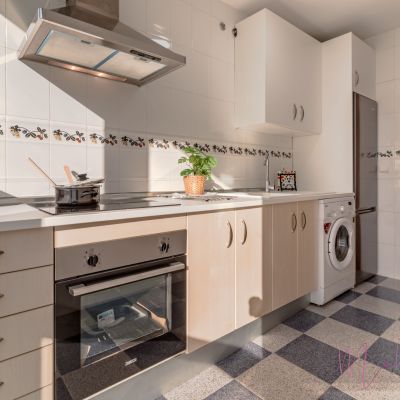 Cocina luminosa con muebles de madera clara, encimera blanca, electrodomésticos de acero inoxidable y azulejos decorativos con detalles florales.