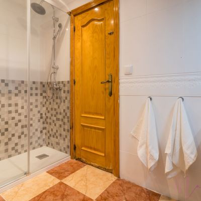 Baño con ducha moderna, mampara de cristal, azulejos en tonos neutros, toallas blancas y puerta de madera.