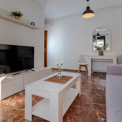 Salón moderno con sofá lila, mesa de centro blanca, televisor, decoración minimalista y suelo de mármol.