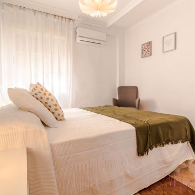 Dormitorio con cama de matrimonio, ropa de cama en tonos blancos y verde oliva, iluminación cálida, aire acondicionado y perchero abierto.