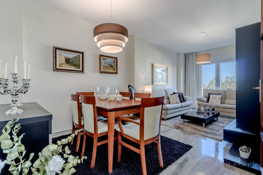 Salón-comedor elegante con mesa de madera, sillas tapizadas en beige, sofás claros con cojines decorativos y una mesa de centro negra. Decoración clásica con cuadros en la pared, candelabro plateado y alfombras en tonos oscuros.