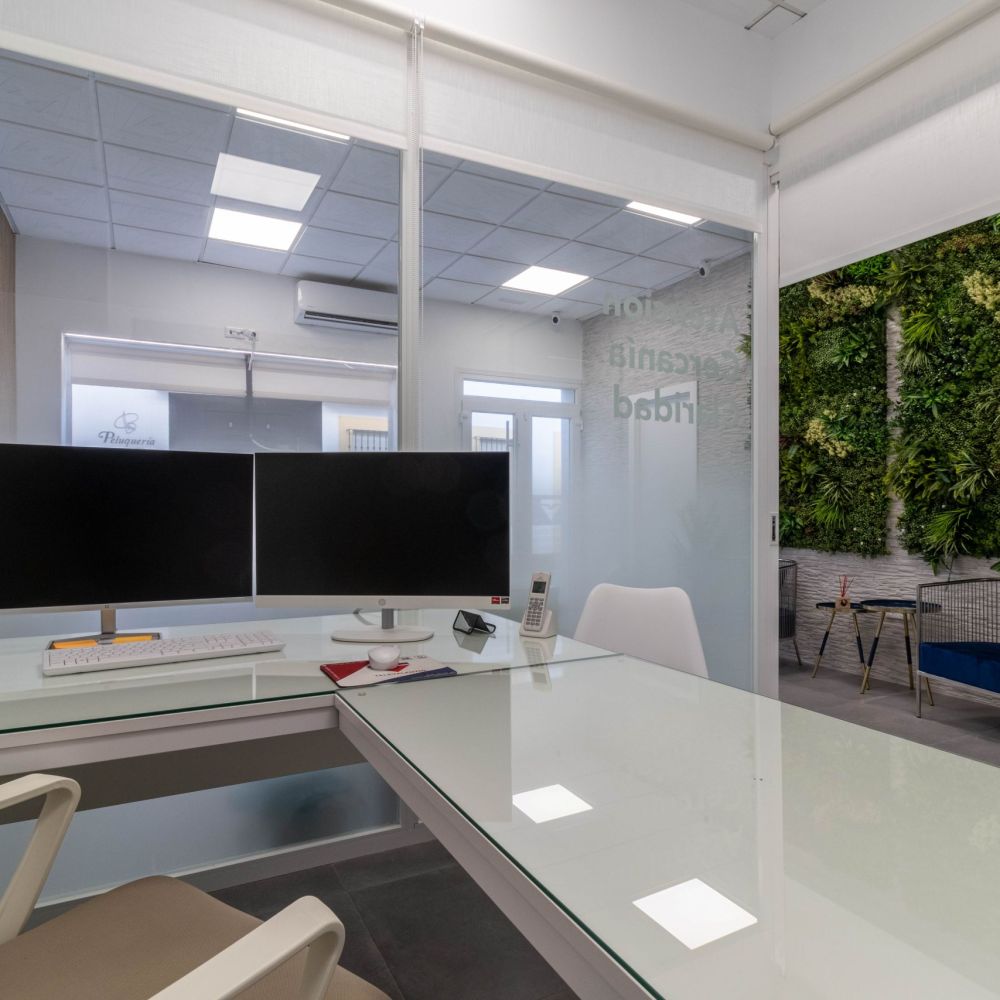 Oficina moderna con escritorio de vidrio, doble monitor, silla ergonómica y separaciones de cristal. Espacio de trabajo profesional con jardín vertical en la sala de reuniones y diseño minimalista.