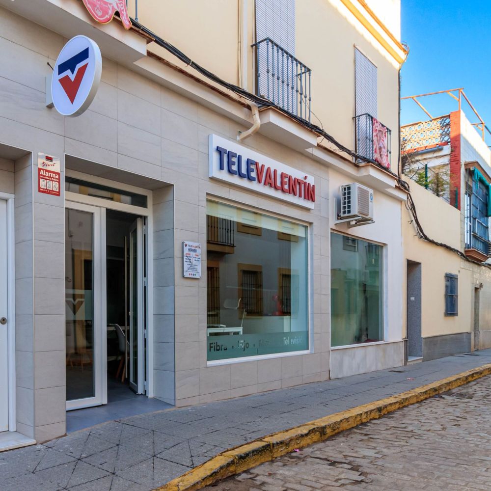 Fachada de TeleValentín, tienda de telecomunicaciones con rótulo en azul y rojo, puerta de entrada acristalada y gran ventanal. Ubicada en una calle adoquinada con edificios tradicionales y cielo despejado