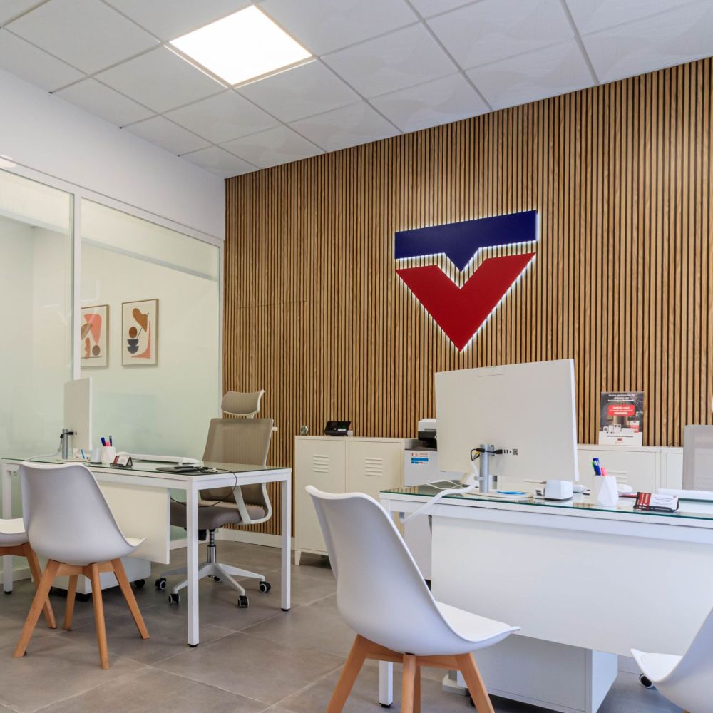 Oficina moderna y elegante con escritorios de vidrio, sillas blancas de diseño y pared decorativa de listones de madera con el logo corporativo iluminado. Espacio minimalista con separaciones de cristal y detalles en tonos neutros