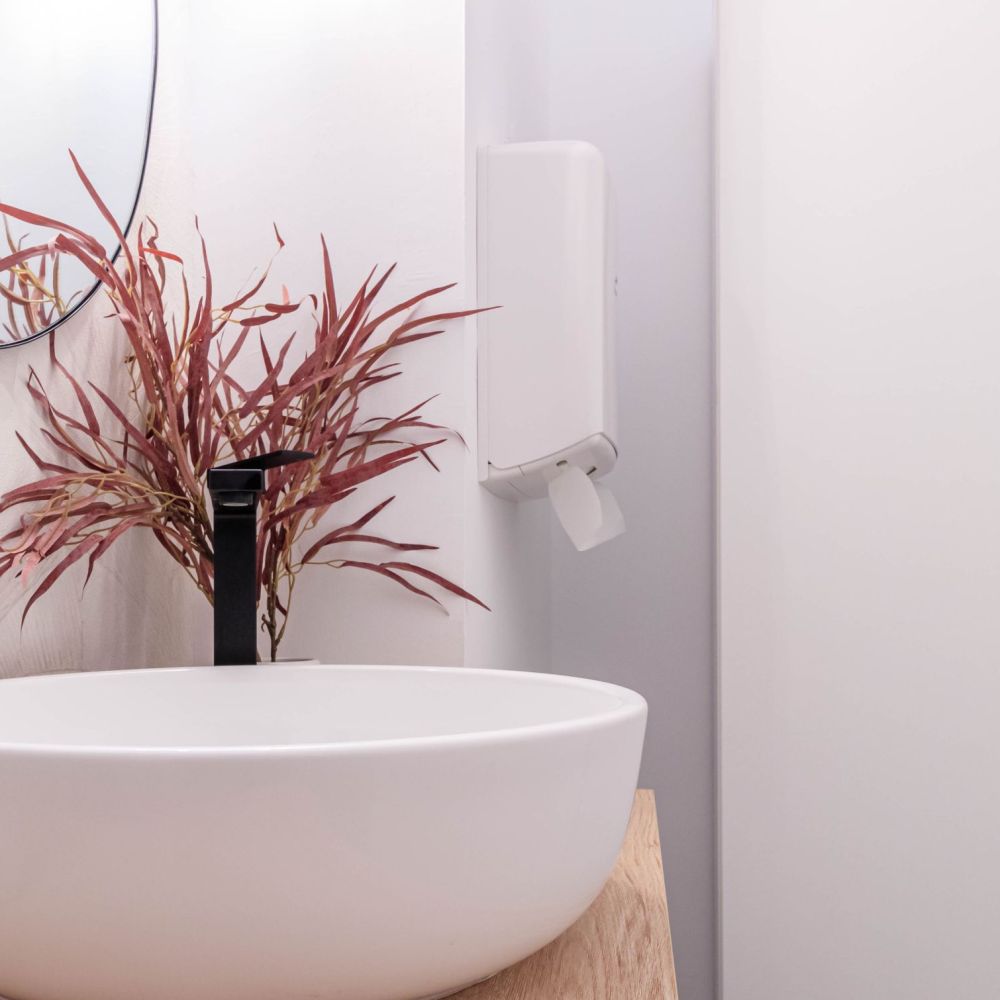 Baño moderno con lavabo sobre encimera de madera, grifo negro y espejo redondo con marco negro. Decoración minimalista con planta seca en tonos rojizos y dispensador de papel en la pared.