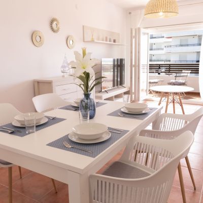 Comedor moderno con mesa blanca, sillas de diseño, vajilla elegante y acceso a un balcón soleado con mobiliario exterior.